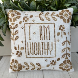 May include: A white pillow with brown embroidery featuring the words "I am worthy" surrounded by floral designs.