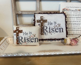 May include: Two white embroidered pillow covers with brown crosses and the text "He is Risen". The pillows are displayed on a white tray with a decorative edge. A sheet of music with the text "I Know That My Redeemer Lives" is visible in the background.