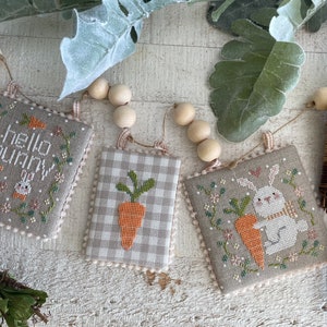 Puede incluir: Tres adornos de tela bordados con un conejito, una zanahoria y el texto "hello bunny". Los adornos están enhebrados con cuentas de madera y cuerda.