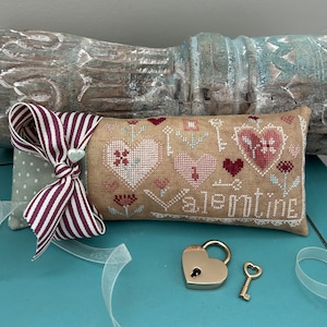 Op de afbeelding: Een beige stoffen kussen met een kruissteekontwerp met harten, bloemen en de tekst "My Valentine". Het kussen is versierd met een wit-burgundy gestreept lint en een witte parelknop. Een hartvormig hangslot en een sleutel liggen op een blauw oppervlak.