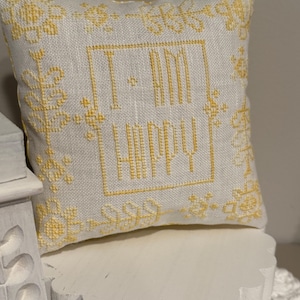 Op de afbeelding: Een klein, vierkant decoratief kussen met de woorden "I AM HAPPY" geborduurd in gele draad. Het kussen heeft een witte achtergrond met gele bloemenborduurwerk rond de randen. Het ligt op een wit, geschulpte plank.