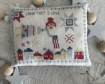 Puede incluir: Una almohada blanca con un diseño de punto de cruz que presenta una bandera estadounidense roja, blanca y azul, una estrella y casas. El texto "Land That I Love" está bordado en hilo negro.
