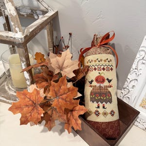 May include: A cylindrical fabric sachet featuring a cross-stitch design of a figure with a pumpkin head, wearing a brown dress and holding two black birds. The top is tied with an orange ribbon, and the base is trimmed with a dark brown floral fabric.