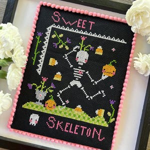 May include: A black cross-stitch embroidery with a pink pom-pom border. The design features a skeleton holding a pumpkin and the text "Sweet Skeleton".
