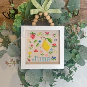 May include: A white framed cross-stitch embroidery with a yellow pear, red strawberries, and colorful flowers. The embroidery says "June" in blue lettering.