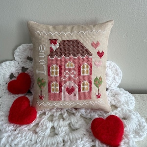 Puede incluir: Un pequeño cojín beige con un diseño de punto de cruz de una casa rosa con tejado marrón, puerta en forma de corazón y la palabra "LOVE". Tres corazones de fieltro rojo y un tapete de ganchillo blanco completan la escena.