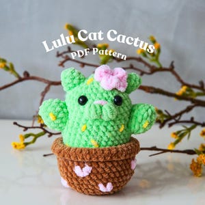 Puede incluir: Un cactus verde tejido a mano con forma de gato, ojos negros y una flor rosa. Está en una maceta marrón con detalles de corazones rosas. El texto "Lulu Cat Cactus PDF Pattern" está arriba. Un artículo hecho a mano.