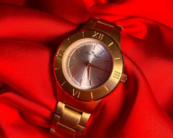 May include: A gold-tone wristwatch with a white face and Roman numeral hour markers. The watch is on a red fabric background.