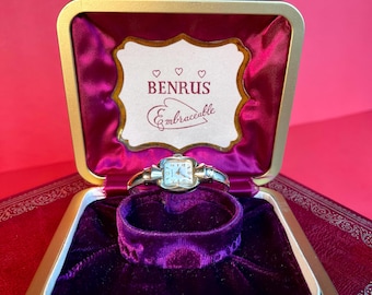 May include: A gold-colored wristwatch with a white face and black Roman numeral hour markers. The watch is in a purple velvet-lined box with a gold-colored label that reads "BENRUS Embracciable" with three hearts.