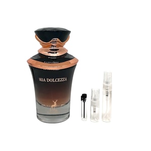 May include: A perfume bottle with a black faceted cap and rose gold accents. The bottle has a gradient brown color and the words "MIA DOLCEZZA" are printed on the front. Several small sample vials are next to the bottle.