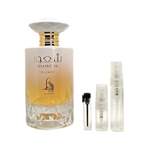 May include: A clear glass bottle of perfume with a gold cap and a white label that says "Shahoor Elixir Al-Agsar". The bottle is surrounded by three small clear glass spray bottles.