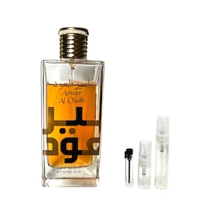 May include: A clear glass bottle of Ameer Al Oudh Intense Oud perfume with a gold cap and label. The bottle is sitting on a white surface with two small clear glass sample bottles with black and clear caps.