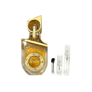 May include: A gold-colored perfume bottle with the word "ZENITH" on the front. The bottle has a unique, angular design and is accompanied by three small sample vials. The perfume bottle and vials are on a white background.