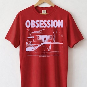 May include: Red t-shirt with the word "OBSESSION" in large, light purple letters across the chest. Below the word is a light purple graphic of a house, car, and person. The shirt is hanging on a wooden hanger.