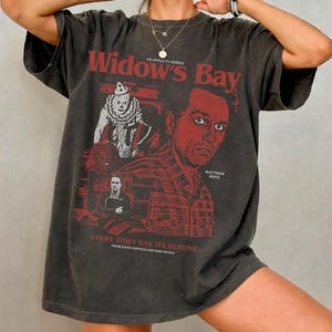 Puede incluir: Una camiseta de manga corta gris carbón con un estampado gráfico rojo titulado Widow's Bay. El diseño incluye un payaso, un hombre y el texto Every Town Has Its Demons, Matthew Rhys, y los nombres de Katie Dippold y Hiro Murai.