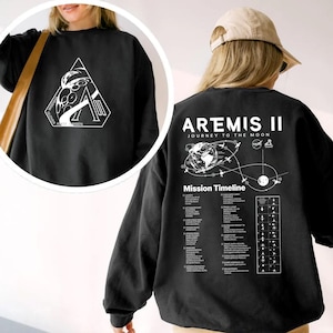 May include: Black sweatshirt with white graphic designs. The front features a geometric design with text, while the back displays "ARTEMIS II JOURNEY TO THE MOON" and a mission timeline. The sweatshirt is a casual garment.