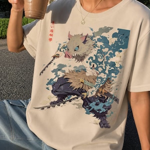 Peut inclure: Un t-shirt beige orné d'une illustration graphique représentant un personnage à tête de sanglier avec des motifs bleus tourbillonnants sur la peau, tenant deux épées. Le design inclut du texte japonais dans un encadré rouge vertical.