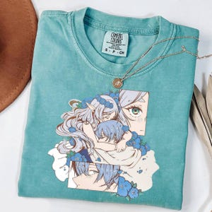 May include: A teal short-sleeve t-shirt featuring a graphic print of two anime-style characters with light blue hair surrounded by blue flowers. The shirt has a Comfort Colors brand label at the neck and a thin gold-tone chain necklace resting on it.
