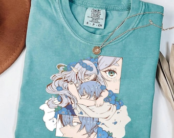 Op de afbeelding: Een blauwgroen t-shirt met korte mouwen en een grafische print van twee anime-stijl personages met lichtblauw haar, omringd door blauwe bloemen. Het shirt heeft een Comfort Colors merklabel in de nek en een dunne goudkleurige ketting.