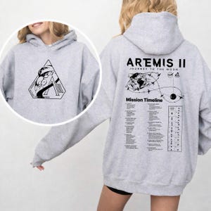 May include: A light gray hoodie featuring a black graphic on the front and back. The front graphic is a triangle with a space-themed design. The back of the hoodie displays the text "ARTEMIS II JOURNEY TO THE MOON" with a mission timeline.