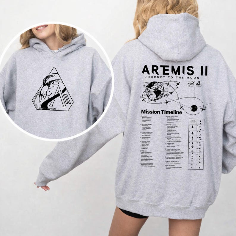 May include: A light gray hoodie featuring a black graphic on the front and back. The front graphic is a triangle with a space-themed design. The back of the hoodie displays the text "ARTEMIS II JOURNEY TO THE MOON" with a mission timeline.