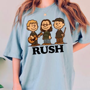 K&ouml;nnte beinhalten: Hellblaues T-Shirt mit einer Cartoon-Illustration von drei Musikern und dem Wort "RUSH" in Schwarz. Die Zeichnung zeigt einen Gitarristen, einen Musiker mit Brille und einen Schlagzeuger mit einem Drumstick, alle im Cartoon-Stil.