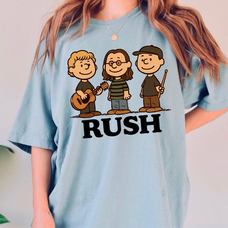 K&ouml;nnte beinhalten: Hellblaues T-Shirt mit einer Cartoon-Illustration von drei Musikern und dem Wort "RUSH" in Schwarz. Die Zeichnung zeigt einen Gitarristen, einen Musiker mit Brille und einen Schlagzeuger mit einem Drumstick, alle im Cartoon-Stil.