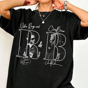 May include: A black short-sleeve t-shirt featuring a white graphic print with the text Usher Raymond and Chris Brown above two black and white portraits. The design includes a large white outline of the letters R and B with signatures below.