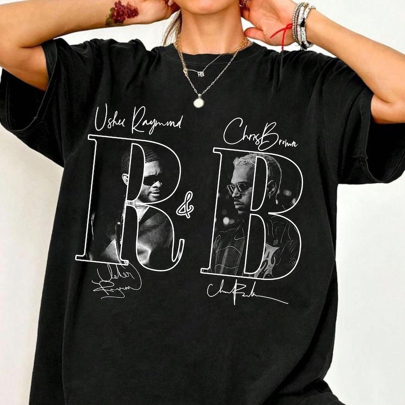 May include: A black short-sleeve t-shirt featuring a white graphic print with the text Usher Raymond and Chris Brown above two black and white portraits. The design includes a large white outline of the letters R and B with signatures below.