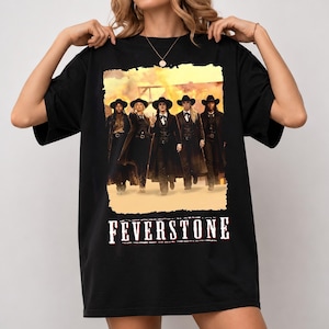 May include: A black short-sleeve t-shirt featuring a graphic print of five individuals in western-style attire, including long coats and cowboy hats, standing in a dusty landscape. The word FEVERSTONE is printed in white below the graphic.