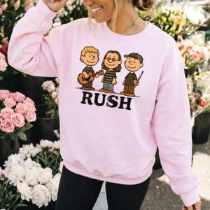 K&ouml;nnte beinhalten: Hellrosa Sweatshirt mit einem Cartoon-Motiv von drei Figuren mit Musikinstrumenten und dem Wort "RUSH" in schwarzen Blockbuchstaben. Die Figuren sind braun, schwarz und gr&uuml;n.