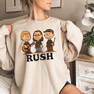 K&ouml;nnte beinhalten: Beiges Sweatshirt mit einer Cartoon-Illustration von drei Figuren, die Musikinstrumente spielen. Die Figuren sind im Stil der Peanuts-Charaktere gehalten. Das Wort "RUSH" ist in schwarzen Blockbuchstaben darunter gedruckt.