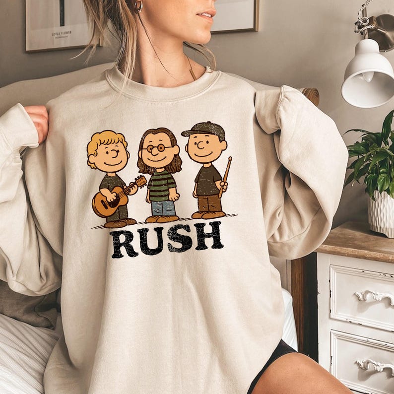 K&ouml;nnte beinhalten: Beiges Sweatshirt mit einer Cartoon-Illustration von drei Figuren, die Musikinstrumente spielen. Die Figuren sind im Stil der Peanuts-Charaktere gehalten. Das Wort "RUSH" ist in schwarzen Blockbuchstaben darunter gedruckt.