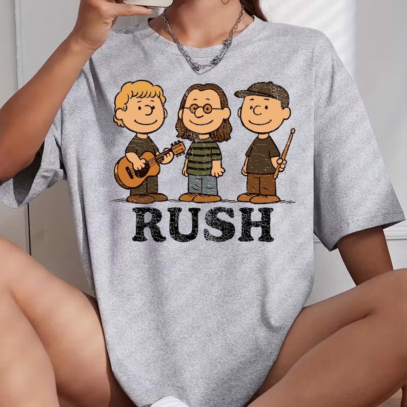 K&ouml;nnte beinhalten: Hellgraues T-Shirt mit einer Cartoon-Illustration von drei Figuren, die Musikinstrumente spielen. Das Wort "RUSH" ist in fetten, schwarzen Buchstaben unter den Figuren aufgedruckt. Die Figuren tragen braune und schwarze Outfits.