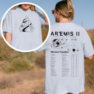 May include: Light blue t-shirt featuring a black graphic on the front and back. The front graphic is a geometric design with text. The back of the shirt displays "Artemis II Journey to the Moon" with a mission timeline and additional text.