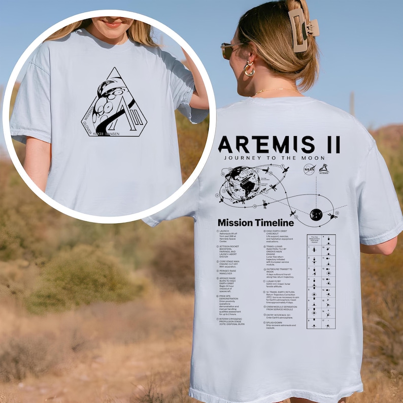 May include: Light blue t-shirt featuring a black graphic on the front and back. The front graphic is a geometric design with text. The back of the shirt displays "Artemis II Journey to the Moon" with a mission timeline and additional text.