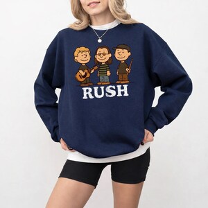 K&ouml;nnte beinhalten: Marineblaues Sweatshirt mit einer Cartoon-Illustration von drei Musikern und dem Wort "RUSH" in Wei&szlig;. Einer spielt Gitarre, ein anderer tr&auml;gt eine Brille und der dritte h&auml;lt einen Drumstick.