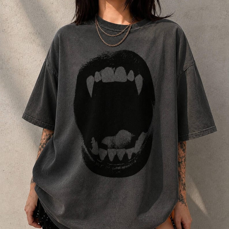May include: A charcoal gray short-sleeve t-shirt featuring a large, black graphic print of an open mouth with prominent fangs. The fabric has a faded, washed-out appearance, and the shirt is styled with a crew neckline and a loose, relaxed fit.