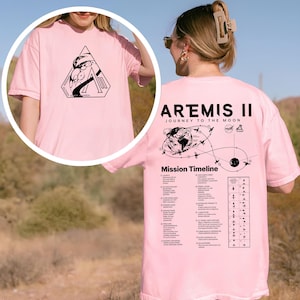 May include: Pink t-shirt featuring a black graphic on the front and back. The front graphic is a geometric design with text. The back of the shirt displays "ARTEMIS II JOURNEY TO THE MOON" and a mission timeline.
