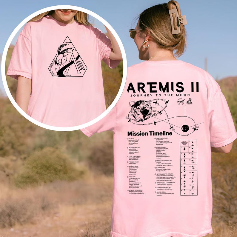 May include: Pink t-shirt featuring a black graphic on the front and back. The front graphic is a geometric design with text. The back of the shirt displays "ARTEMIS II JOURNEY TO THE MOON" and a mission timeline.