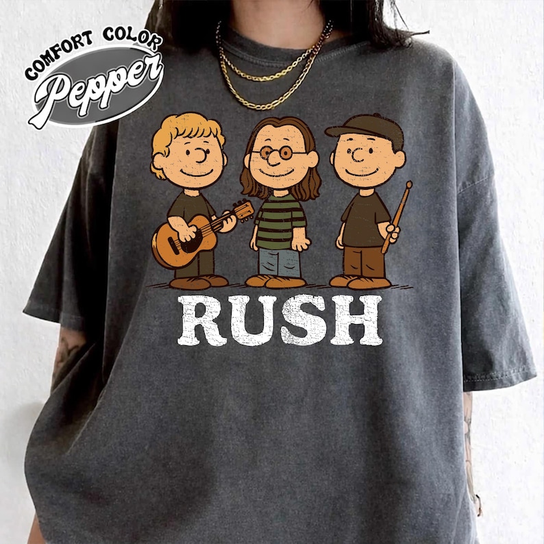 K&ouml;nnte beinhalten: Dunkelgraues T-Shirt mit einer Cartoon-Illustration von drei Musikern. Das Wort "RUSH" ist in wei&szlig;en Blockbuchstaben unter den Figuren aufgedruckt. Das Shirt hat auch die Worte "Comfort Color Pepper" in einem wei&szlig;en Oval aufgedruckt.