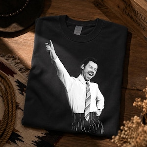 May include: A black crewneck t-shirt featuring a monochromatic graphic print of a person with a mustache wearing a button-down shirt, a striped necktie, and pinstriped trousers, with one arm raised while holding a microphone in a performance pose.