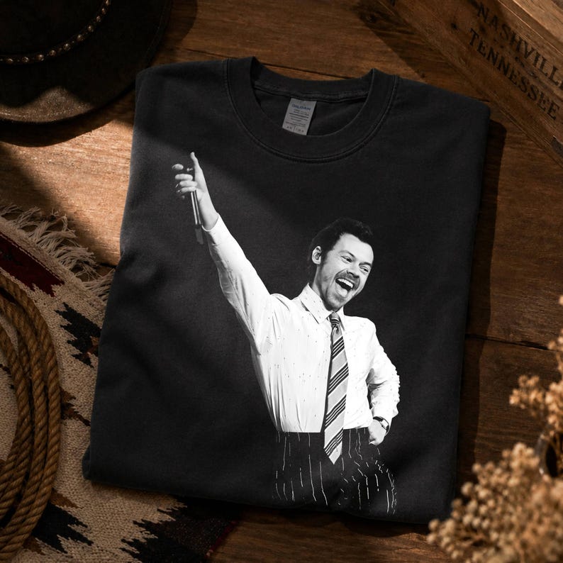 May include: A black crewneck t-shirt featuring a monochromatic graphic print of a person with a mustache wearing a button-down shirt, a striped necktie, and pinstriped trousers, with one arm raised while holding a microphone in a performance pose.