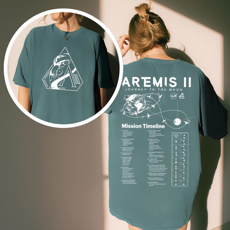 May include: Teal-colored t-shirt featuring a white graphic on the front and back. The front graphic is a triangle with a space-themed design and the text "KAZI HANSEN". The back of the shirt displays "ARTEMIS II JOURNEY TO THE MOON" and a mission timeline.