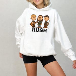 K&ouml;nnte beinhalten: Wei&szlig;er Kapuzenpullover mit einer Cartoon-Illustration von drei Musikern und dem Wort "RUSH" in schwarzem Text. Die Musiker spielen Instrumente, darunter eine Gitarre und Trommelst&ouml;cke. Der Hoodie hat eine Vordertasche und eine Kapuze mit Kordelzug.
