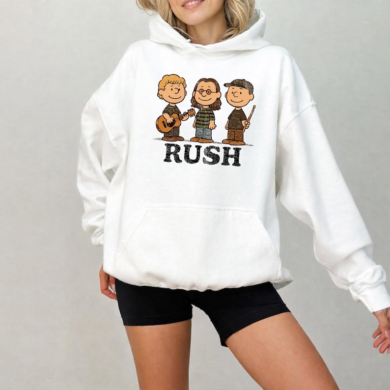 K&ouml;nnte beinhalten: Wei&szlig;er Kapuzenpullover mit einer Cartoon-Illustration von drei Musikern und dem Wort "RUSH" in schwarzem Text. Die Musiker spielen Instrumente, darunter eine Gitarre und Trommelst&ouml;cke. Der Hoodie hat eine Vordertasche und eine Kapuze mit Kordelzug.