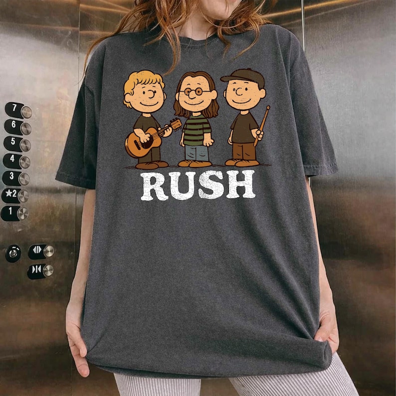 K&ouml;nnte beinhalten: Dunkelgraues T-Shirt mit einer Cartoon-Illustration von drei Musikern und dem Wort "RUSH" in Wei&szlig;. Eine Figur h&auml;lt eine Gitarre, eine andere tr&auml;gt eine Brille und ein gestreiftes Hemd, die dritte h&auml;lt Trommelst&ouml;cke.