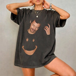 May include: A charcoal gray short-sleeve t-shirt featuring a graphic print of a man's face positioned above a large, curved, smiling mouth shape. The shirt has a relaxed fit, a crew neckline, and a faded, vintage-style wash throughout the fabric.
