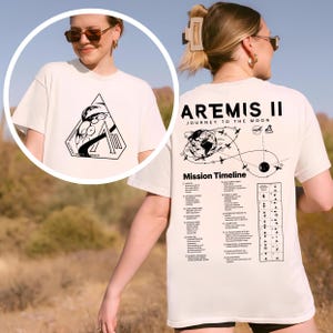 May include: Cream-colored t-shirt featuring a black graphic on the front with the text "WISDOM GLOVER KAZI HANSEN" and a large letter "A". The back of the shirt displays "ARTEMIS II JOURNEY TO THE MOON" with a mission timeline graphic.