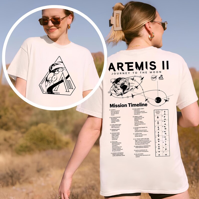 May include: Cream-colored t-shirt featuring a black graphic on the front with the text "WISDOM GLOVER KAZI HANSEN" and a large letter "A". The back of the shirt displays "ARTEMIS II JOURNEY TO THE MOON" with a mission timeline graphic.